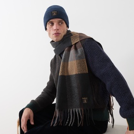 Model wearing Barbour Westerdale Logo Badge Beanie & Scarf Set in navy/grey; tartan scarf with tasselled ends, fine knit beanie with logo patch