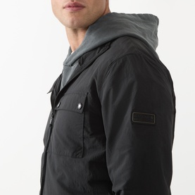 Model wearing Barbour International Dalliston Logo Jacket in black, showing logo sleeve patch, chest pocket, and grey hood detail.