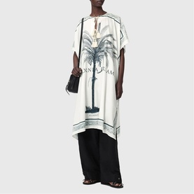 Model wearing All Saints Tess Printed Logo Kaftan in ecru with palm tree print, tassel ties, styled with black wide-leg trousers and sandals.