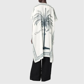 Model wearing All Saints Tess Printed Logo Kaftan in ecru white with palm tree print, styled with black wide-leg trousers.