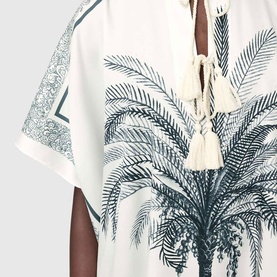 Model wearing All Saints kaftan in ecru white with teal palm tree print, tassel tie neckline, and short sleeves. Close-up detail shot.