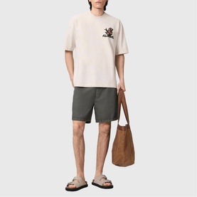 Model wearing All Saints Cacti Oversized Graphic Logo T-Shirt in Dusk White with khaki shorts, styled with brown tote bag and sandals.