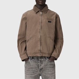 Model wearing All Saints Korey Canvas Jacket in Mire Brown with zip closure, chest pocket, and leather branding detail, styled with grey jeans.