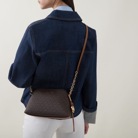 Model carrying MMK Freddy Chain Crossbody Bag in brown logo print with gold-toned hardware and tan leather crossbody strap, styled with denim jacket