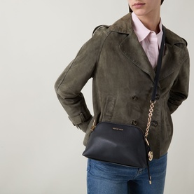 Model carrying MMK Freddy Chain Crossbody Bag in black with gold-toned chain strap and logo hardware, styled with olive suede jacket and jeans