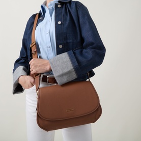 Model carrying MMK Holmes Logo Crossbody Bag in Luggage tan leather, with adjustable crossbody strap and gold-tone hardware, styled with denim jacket