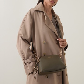 Model carrying MMK Freddie Crossbody Bag in loden, dome silhouette with gold-toned chain strap and monogram keychain, styled with beige trench coat