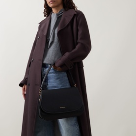 Model carrying MMK Holmes Logo Crossbody Bag in black leather, flap closure with gold logo hardware, worn crossbody over dark coat and jeans.