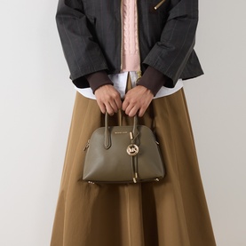 Model carrying MMK Freddie Logo Satchel Bag in loden pebble leather, gold-toned hardware, top handles, styled with olive midi skirt and grey jacket