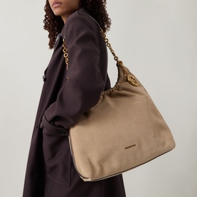 Model carrying MMK Indie Logo Shoulder Bag in khaki leather, gold-toned chain strap and logo detail, styled with dark overcoat.