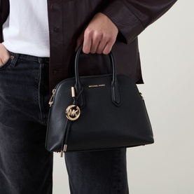 Model carrying MMK Freddie Logo Satchel Bag in black grained leather with gold-tone hardware and branded keychain, paired with grey jeans