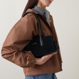 Model carrying MMK Laila Tote Bag in black suede with gold-toned hardware and top handles, styled with a tan jacket.