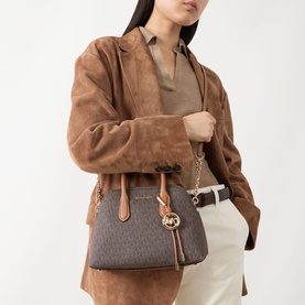 Model carrying MMK Freddie Logo Satchel Bag in brown/acorn with gold-tone chain strap and logo charm, styled with tan suede jacket