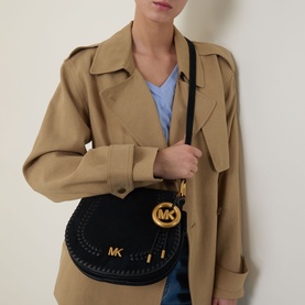 Model carrying MMK Josie Saddle Crossbody Bag in black suede with gold-tone MK logo hardware and braided trim, worn over camel trench coat.