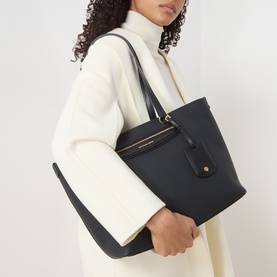 Model carrying MMK Jet Set East West Tote Bag in black grained leather with gold-tone hardware and front zip pocket, styled with cream coat