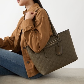 Model carrying MMK Jet Set East West Saffiano Tote in Loden with gold-tone hardware and front zip pocket, styled with brown jacket and jeans