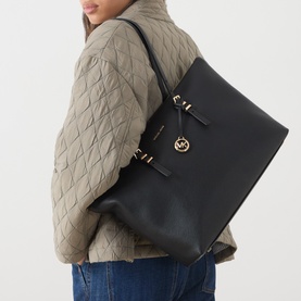Model carrying MMK Quinn Large Tote Bag in black leather with gold-tone hardware and hanging MK logo charm, styled with quilted jacket and jeans