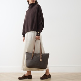 Model carrying MMK Quinn Large Tote Bag in brown monogram canvas with tan straps and gold-tone hardware, styled with dark knitwear and cream skirt.