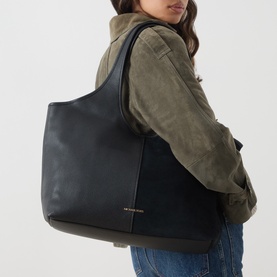 Model carrying MMK Henly Shopper Tote Bag in black leather with gold-tone finishes, worn on shoulder. Styled with khaki jacket and jeans.