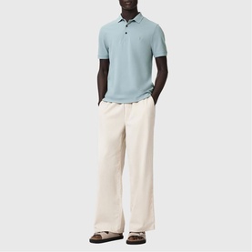 Model wearing All Saints Reform Ramskull Logo Polo Shirt in Salton Blue with cream wide-leg trousers and brown sandals.
