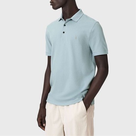 Model wearing All Saints Reform Ramskull Logo Polo Shirt in Salton Blue, styled with cream trousers. Short sleeves, button placket.