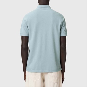 Model wearing All Saints Reform Ramskull Logo Polo Shirt in Salton Blue, back view, styled with off-white trousers.