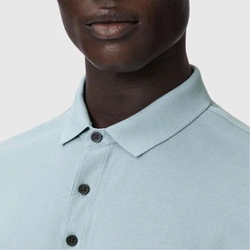 Model wearing All Saints Reform Ramskull Logo Polo Shirt in Salton Blue, close-up of polo collar and three-button placket detail.