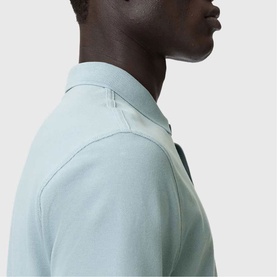 Model wearing All Saints Reform Ramskull Logo Polo Shirt in Salton Blue, close-up showing polo collar and pique fabric detail.