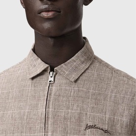 Model wearing All Saints Keal Check Jacket in sediment brown, close-up of zip fastening and chainstitch embroidery logo detail.