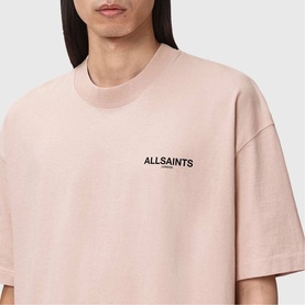 Model wearing AllSaints Hallis Oversized Graphic Logo T-Shirt in Sunbaked Pink with front logo print, crew neck, relaxed fit.
