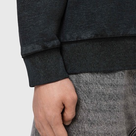 Model wearing All Saints washed black sweatshirt, close-up of ribbed cuff detail, styled with grey marl trousers.