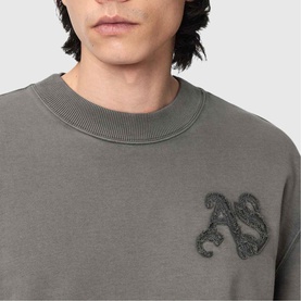 Model wearing All Saints Insignia Logo Sweatshirt in Washed Black with gothic appliqu&eacute; logo detail on chest, crew neckline with ribbed trim.