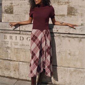 Model wearing Mint Velvet red check midi skirt styled with a burgundy short-sleeve ribbed top, asymmetric hem, outdoor setting