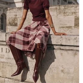 Model wearing Mint Velvet red plaid midi skirt, styled with burgundy knit top and knee-high leather boots, seated on stone ledge.