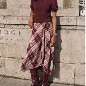 Model wearing Mint Velvet red plaid asymmetric midi skirt with burgundy knit top and dark brown knee-high leather boots.
