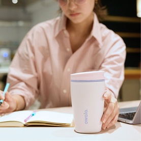 Model with Owala SmoothSip&reg; Slider Travel Mug in Candy Coated, pale pink with purple accent lid, held at desk while writing