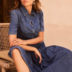 Model wearing Mint Velvet blue denim midi dress with zip front, collar, and short sleeves, seated, styled with gold hoop earrings.