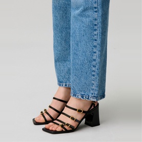 Model wearing Claude Pierlot black block-heel sandals with gold-tone buckle straps, styled with light blue wide-leg jeans