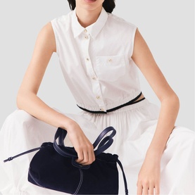 Model carrying Maje metallic disc bag in navy blue with drawstring closure and suede-effect finish, styled with white sleeveless shirt and skirt