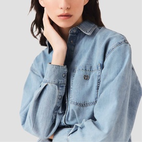 Model wearing Maje light blue denim shirt with chest pocket and small logo pin detail, cropped close-up shot