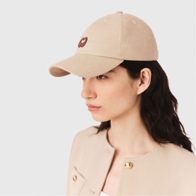 Model with Maje baseball cap in clear beige, small logo patch to front, worn with a beige coat