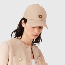 Model wearing Maje baseball cap in clear beige with brown heart logo embroidery, styled with a matching beige jacket with gold buttons