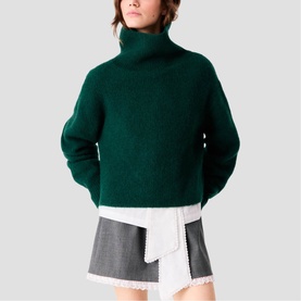 Model wearing Maje bottle green chunky knit turtleneck sweater, styled with a grey mini skirt and white shirt layer.