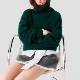Model wearing Maje bottle green chunky turtleneck sweater, arms crossed, styled with a grey frayed mini skirt