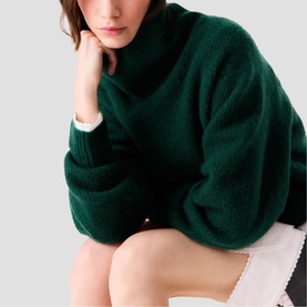 Model wearing Maje bottle green chunky turtleneck sweater with oversized fit and balloon sleeves, styled with white shorts