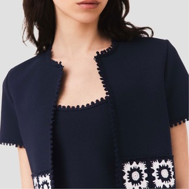 Model wearing Maje navy short-sleeve jacket with white crochet floral trim and scalloped edges, styled over matching navy dress.