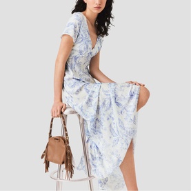 Model wearing Maje Womens French Dress in sky blue large floral print, short sleeves, V-neck, midi length. Styled with tan fringed bag.