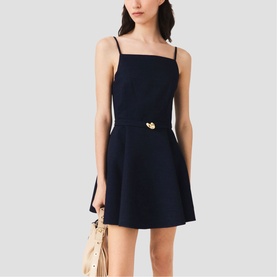 Model wearing Maje Womens French Dress in navy, featuring thin straps, gold heart belt detail and A-line skirt, styled with a woven bag.