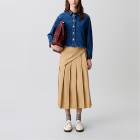 Model wearing Claude Pierlot Pleated Drape Skirt in clear tan, midi-length with side pleats, styled with denim shirt and carrying burgundy clutch.