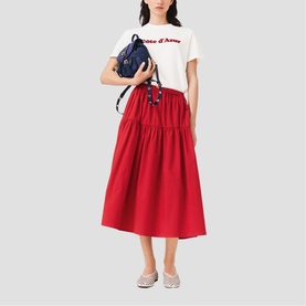 Model wearing Maje red midi skirt with gathered waist, styled with white slogan tee, carrying navy backpack, wearing grey trainers